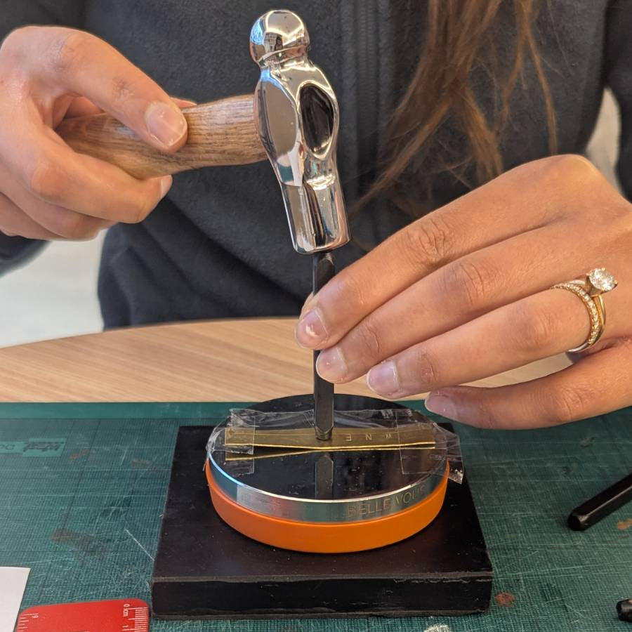 Person using a hammer on a small metal object with tools and a ruler on a workbench.